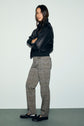 A woman stands with arms crossed, wearing a black jacket, a motif pied-de-poule pantalon slim longueur 7/8, and black loafers, looking at the camera against a plain light background.