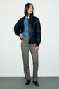 Woman stands against a plain background, wearing a black leather jacket, blue sweater, and a pantalon slim à motif pied-de-poule de longueur 7/8 paired with black loafers. She has long dark hair and her left hand rests on her hip while she faces forward.