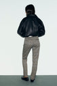 A person stands facing away, wearing a black leather jacket, slim-fit pantalon motif pied-de-poule à longueur 7/8, and black shoes, against a plain light background.