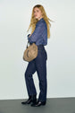 A person with long blonde hair wearing a blue shirt, dark blue jean droit, black boots, and holding a tan shoulder bag stands against a plain light background.