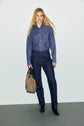 A woman with long blonde hair wears a blue denim shirt, a pair of jean droit in a dark wash, and black boots. She holds a tan handbag in her left hand and stands against a plain, light-colored background.