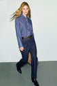 A person with long blonde hair wears a blue denim shirt tucked into a jean brut with a black belt and black boots, walking confidently against a plain light background.