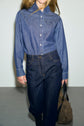 A person in a blue denim button-up shirt and dark jean brut holds a taupe handbag, standing against a plain light background.