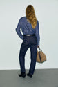 Person with long, wavy hair facing away, wearing a denim shirt, dark jean brut coupe droite, black boots, and holding a beige handbag against a plain background.