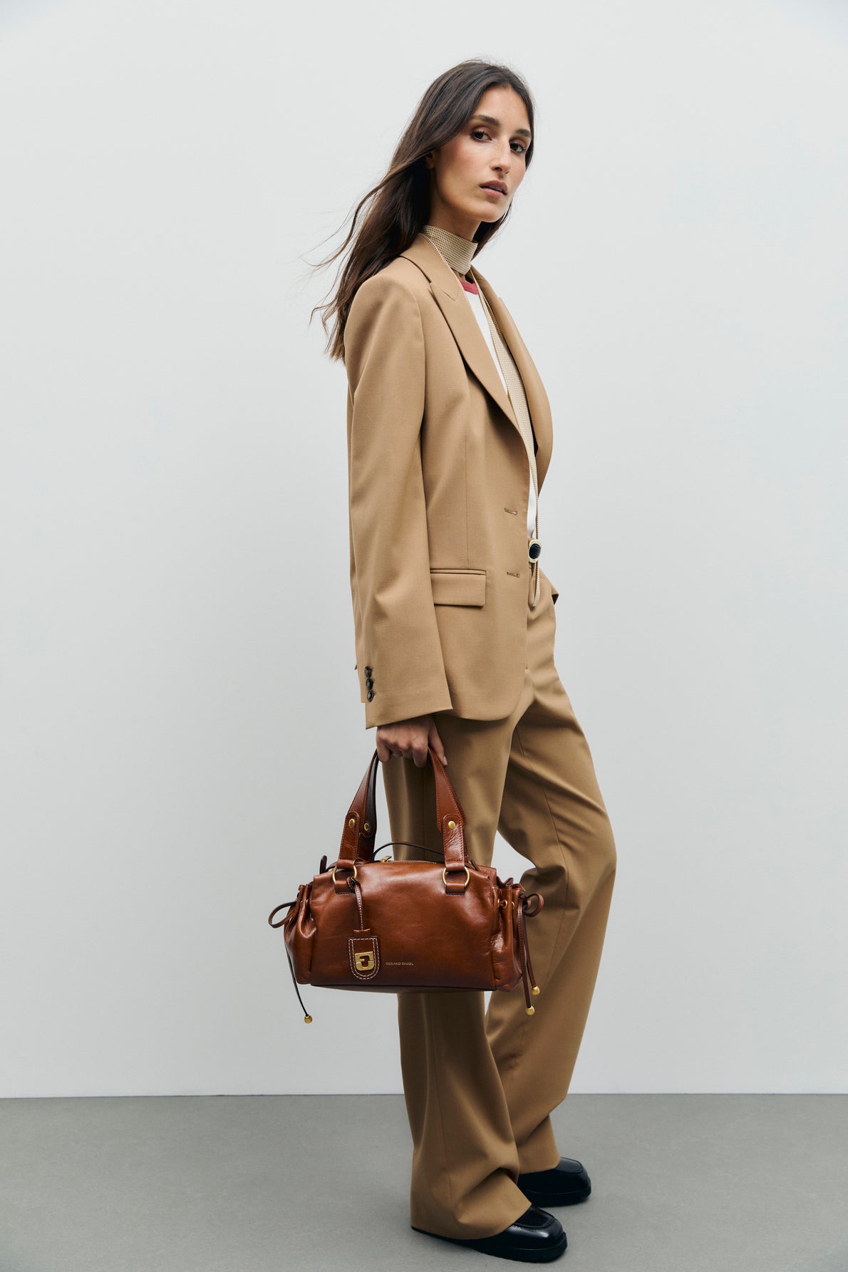 A woman in a tan suit with a pantalon tailleur and white top stands against a plain background, holding a brown leather handbag in her right hand. She has long brown hair and wears black shoes.
