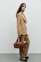 A woman in a tan suit with a pantalon tailleur and white top stands against a plain background, holding a brown leather handbag in her right hand. She has long brown hair and wears black shoes.