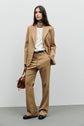 A woman stands against a plain background wearing a pantalon tailleur camel suit, white top, and black shoes. She holds a brown handbag and has long, loose hair, with a neutral expression.