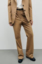 A person wearing a tan suit with a white shirt, black belt, and black shoes stands against a plain gray background. The outfit features a pantalon tailleur camel, with only the body from shoulders down visible.