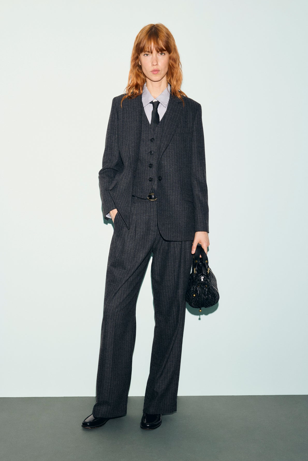 Person with shoulder-length hair wears a dark pinstripe suit with a pantalon rayé, blue shirt, and black tie, holding a black bag. They stand against a plain light background, looking at the camera with one hand in their pocket.