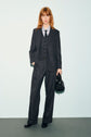 Person with shoulder-length hair wears a dark pinstripe suit with a pantalon rayé, blue shirt, and black tie, holding a black bag. They stand against a plain light background, looking at the camera with one hand in their pocket.