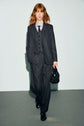 A person with shoulder-length red hair wears a dark pinstripe suit with a flanelle anthracite pantalon à pinces, buttoned vest, tie, and holds a black bag while walking on a gray floor against a pale background.