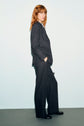 A person with shoulder-length reddish hair stands sideways, wearing a dark flanelle anthracite pinstripe suit and black shoes, hands in pockets, against a plain light background.