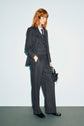 A person with medium-length red hair wears a gray flanelle anthracite pinstripe suit, vest, light blue shirt, and black shoes, standing against a pale background and holding a small black handbag.