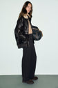 A woman stands against a plain background, wearing a black leather jacket, beige sweater, and a chic pantalon femme—black wide-leg pants that add an elegant touch to her look—paired with brown shoes. She carries a large black patterned handbag with tan handles on her shoulder.