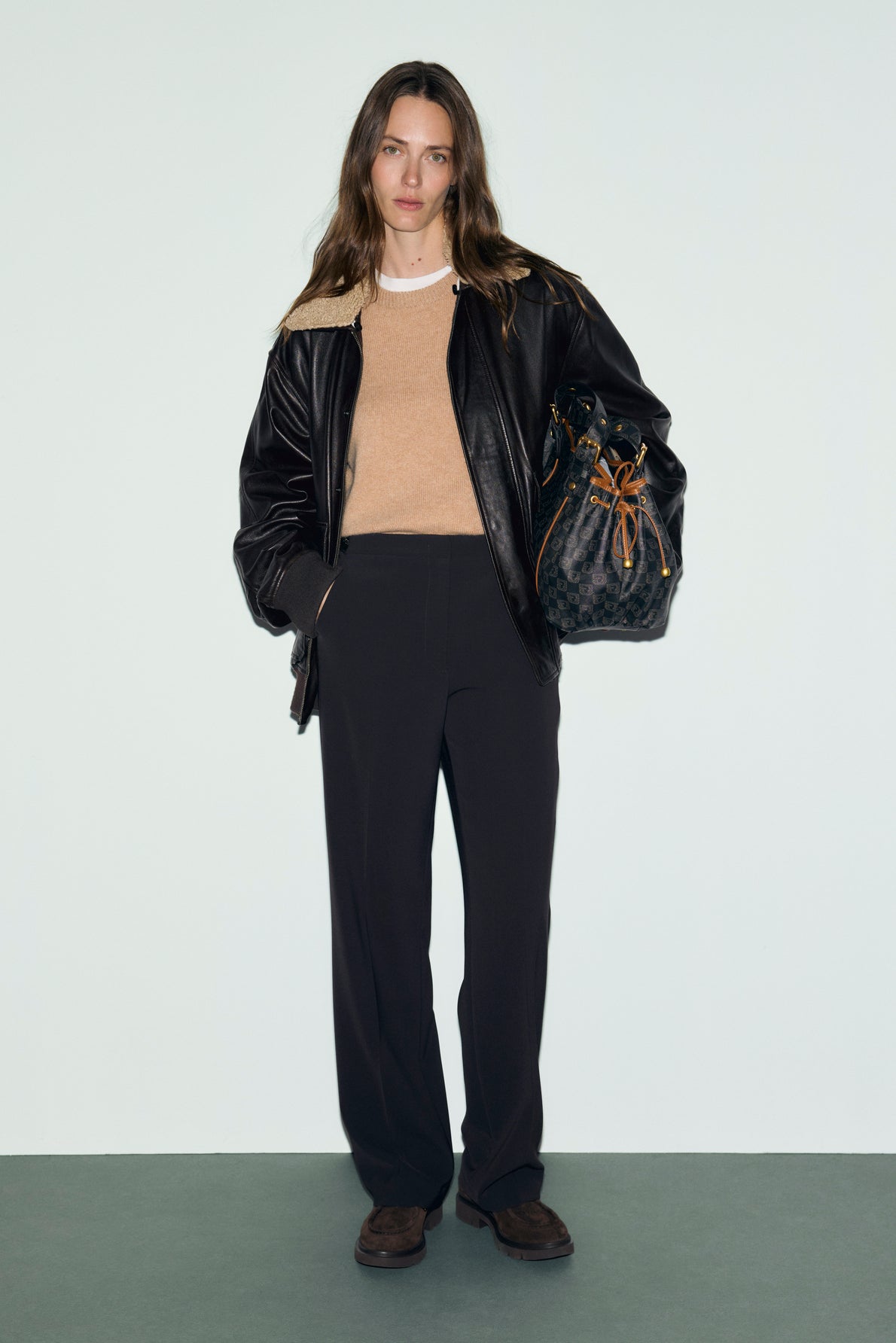 Woman stands against a plain background wearing a black leather jacket with shearling collar, tan sweater, pantalon femme chic in black, and brown shoes. She carries a black bucket bag with tan drawstring.