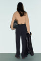 A person with long brown hair, wearing a tan sweater and a pantalon femme chic noir, stands facing away, holding a black bag and a black jacket against a plain light background.