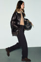 A woman in a black leather jacket, tan sweater, and chic pantalon de tailleur walks while carrying a large black patterned handbag. She has long brown hair and is wearing dark shoes.