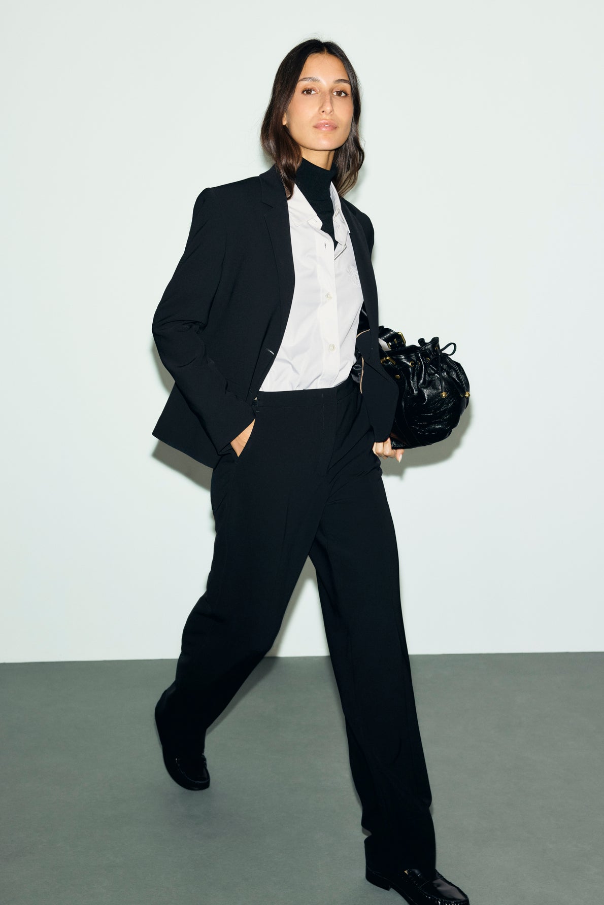 A person in a black suit and white shirt walks forward, holding a black handbag. The outfit features a pantalon droit, adding an elegant touch against the plain, light-colored background.
