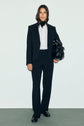 A woman stands against a plain background, wearing a black pantalon de tailleur with a white shirt and black turtleneck. She holds a large black bag and wears black loafers. Her hair is loose and she has a neutral expression.