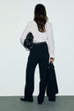 A person with long brown hair, seen from behind, wears a white button-down shirt and a pantalon noir femme de tailleur with a straight cut, holding a black bag under one arm and a black jacket in the other hand, standing against a plain background.
