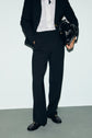 A person stands against a white wall, wearing a black blazer, white button-down shirt, and a pantalon noir femme. They are holding a shiny black handbag in their left hand and wearing black loafers.