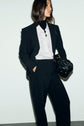 A woman in a black suit and white shirt stands against a plain background, holding a black handbag. She has long brown hair and is looking to the side with one hand in the pocket of her pantalon de tailleur.