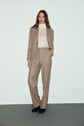 A person with long hair wears a tan suit featuring a pantalon de tailleur in flanelle beige with a coupe droite, paired with a light turtleneck and black shoes, standing against a plain light background.