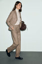 A woman in a tan suit with a coupe droite and white turtleneck walks while holding a brown handbag. She has long brown hair, wears black loafers, and her pantalon de tailleur in flanelle beige adds an elegant touch. The background is plain and light-colored.