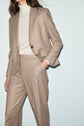 A woman wearing a beige flanelle beige suit with a single-button blazer and matching pantalon de tailleur coupe droite, paired with a light turtleneck sweater, poses against a plain light background. Her hand is in her pocket.