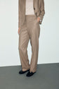Person wearing a tan suit with wide-leg trousers in flanelle beige, a light beige top, and black shoes, standing against a plain light background. The person’s face is not visible in the image.
