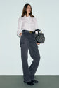 A person stands against a plain background wearing a white blouse, high-waisted gray jean cargo pants à coupe droite with poches plaquées, black shoes, and holding a patterned handbag.