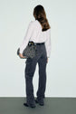 A person with long brown hair, wearing a white blouse and dark gray jean cargo pants with poches plaquées, stands facing away, holding a patterned bag on their left side against a plain background.