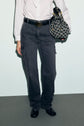 A person wearing a white shirt, dark jean cargo à coupe droite with poches plaquées, a black belt, and black shoes holds a patterned handbag with gold accents. The background is plain and minimal.