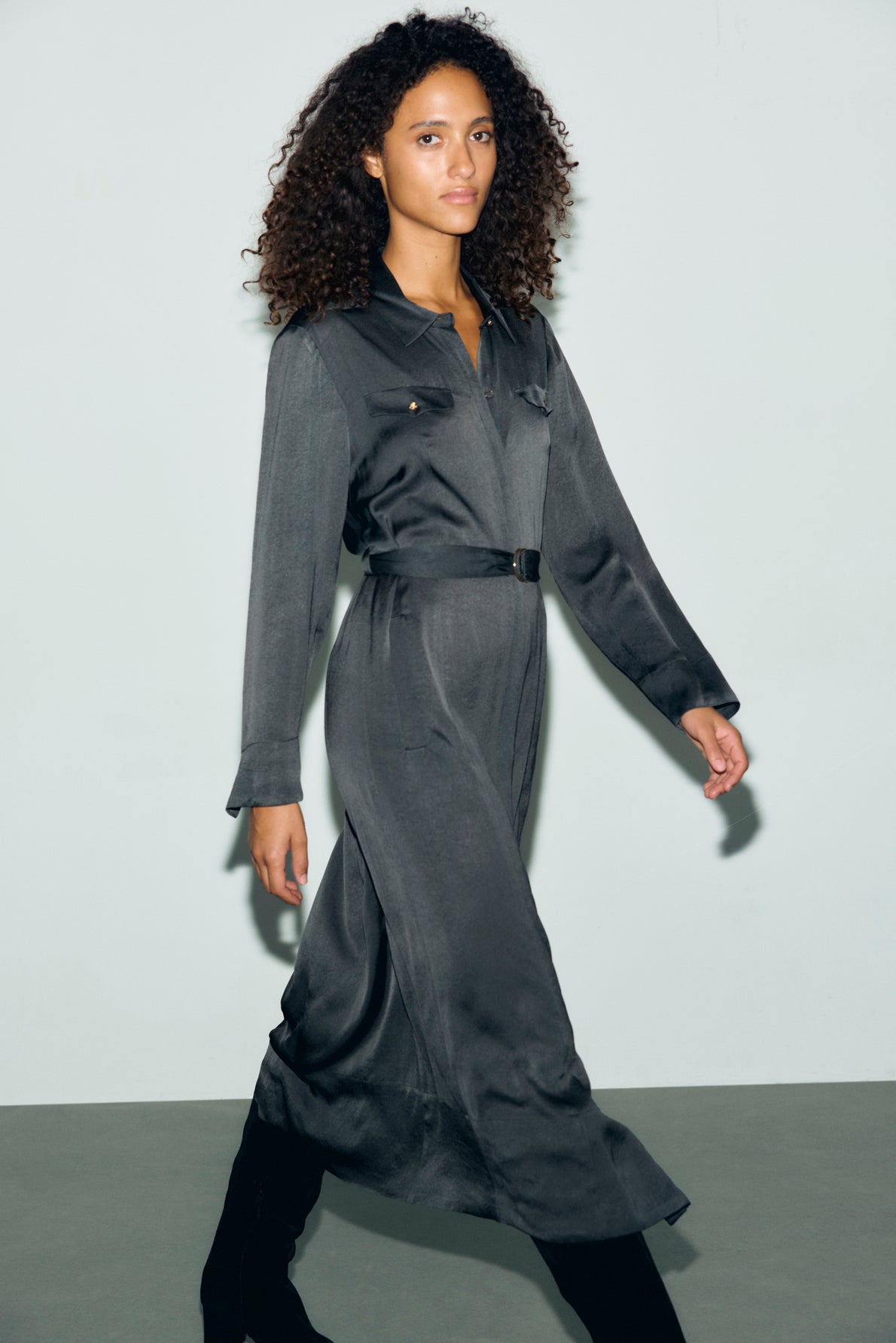 A woman with curly hair walks confidently, wearing a long-sleeved robe midi belted dress in dark gray and black boots, against a plain light background.