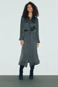 A woman with curly hair wears a long, dark belted coat styled over a robe midi and black boots, standing against a plain, light background.