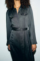 A person with curly hair wears a long-sleeved, belted, black robe chemise satin with buttoned chest pockets, standing against a plain light background. Their face is partially out of frame.