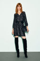 A woman with red hair wears a robe courte à motif imprimé noir à pois blancs, dotée d’un col V élégant et cintrée par une ceinture noire. Elle complète sa tenue avec des bottes noires montant aux genoux et un sac à main noir, se tenant devant un fond clair.