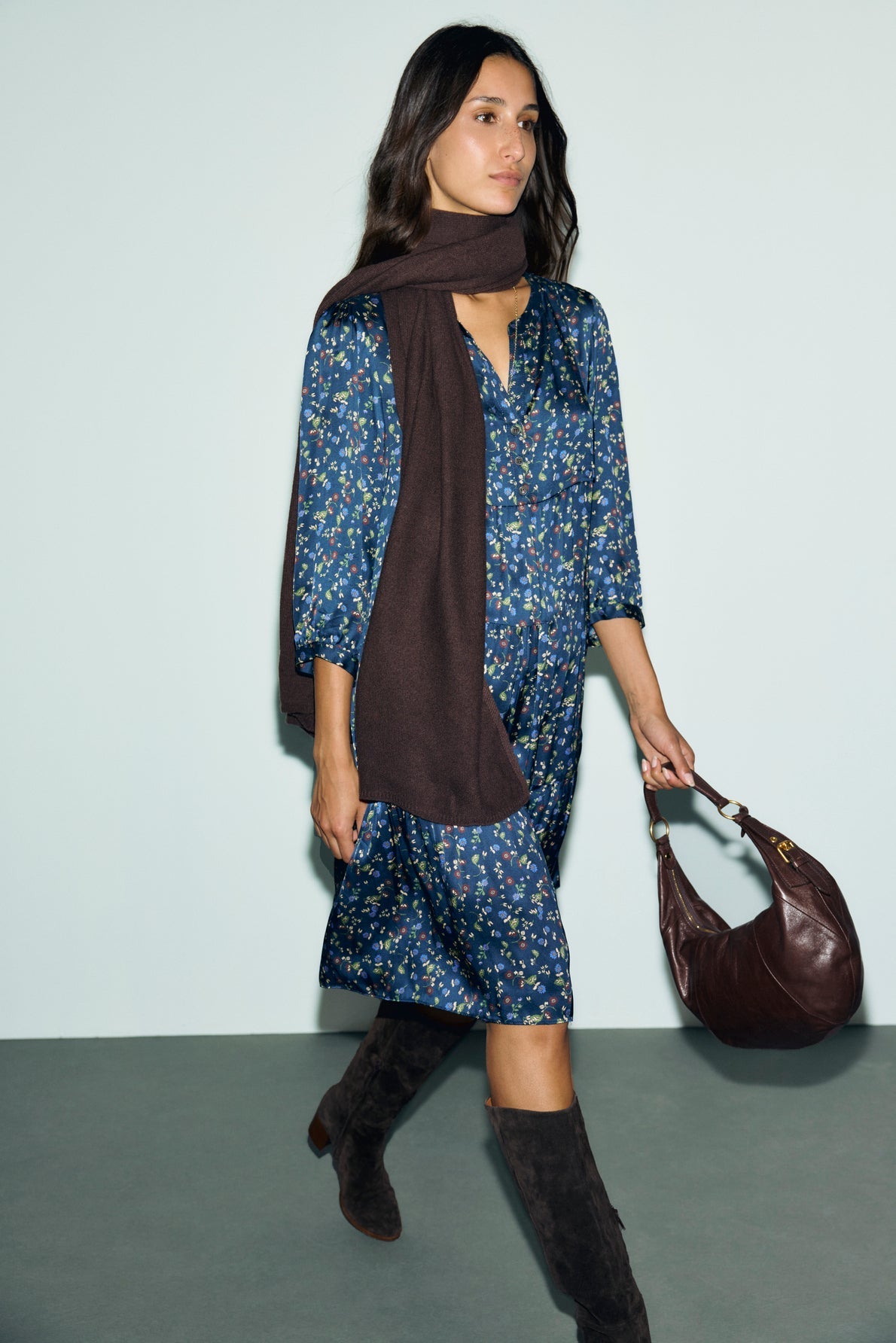 Woman in a blue robe fleurie, brown scarf, and knee-high brown boots walks while carrying a brown shoulder bag against a light background.