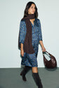 Woman in a blue robe fleurie, brown scarf, and knee-high brown boots walks while carrying a brown shoulder bag against a light background.