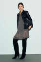 Woman in a black leather jacket, black and white polka dot robe courte with a col chemise, over-the-knee black boots, and holding a black fringed bag stands against a light background.