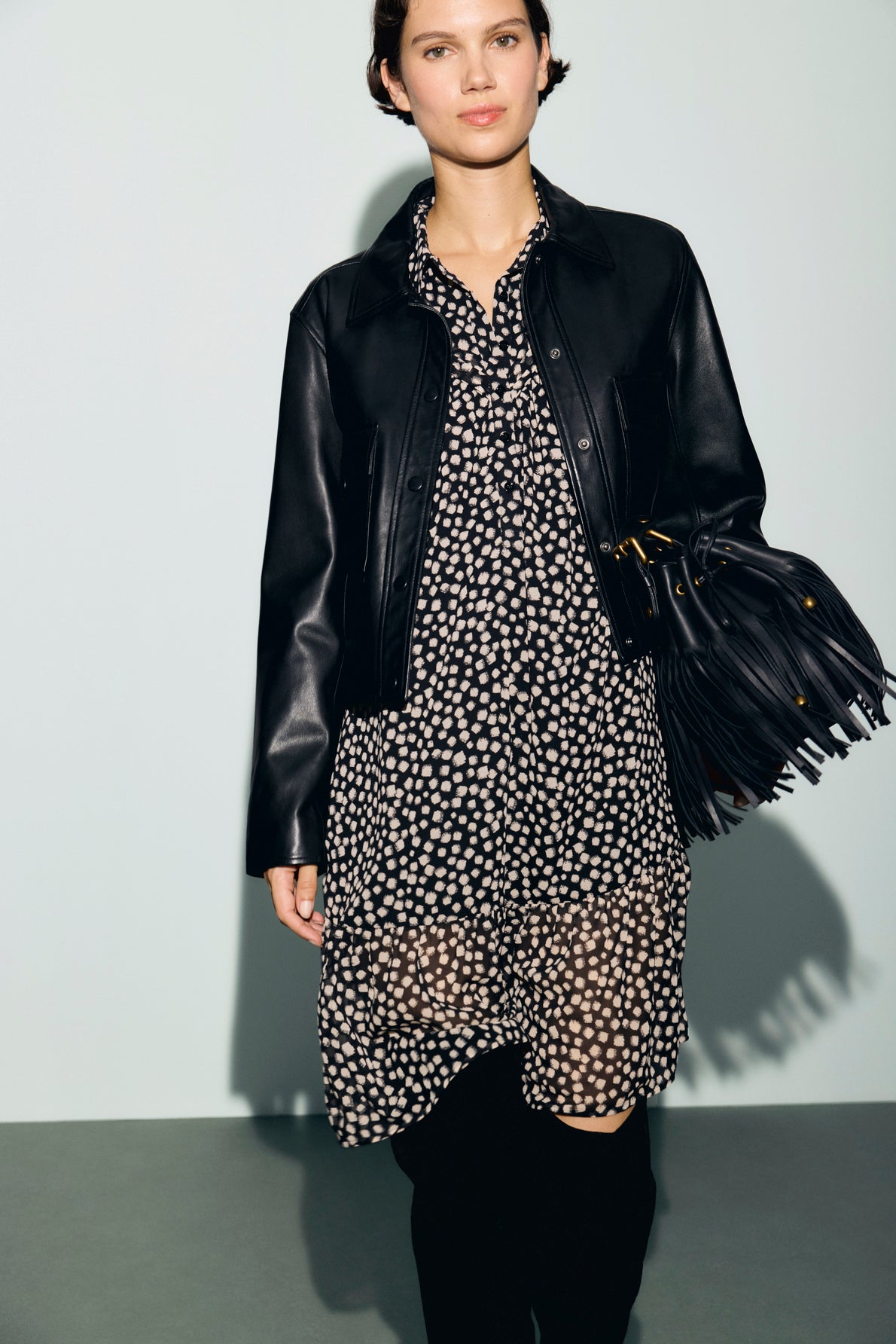 A person wearing a black leather jacket over a black and white polka dot robe courte with col chemise holds a black fringed handbag. They are standing against a plain light background.