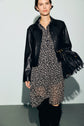 A person wearing a black leather jacket over a black and white polka dot robe courte with col chemise holds a black fringed handbag. They are standing against a plain light background.