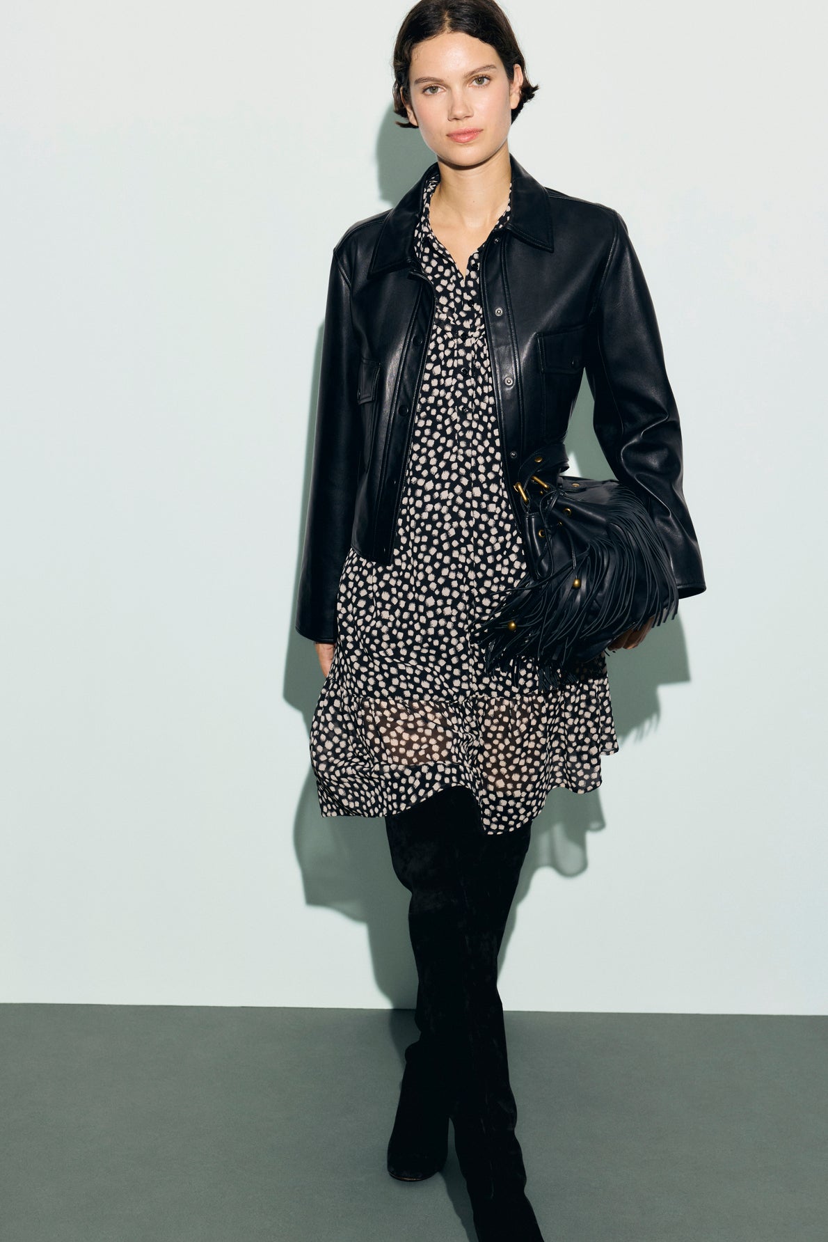 Woman wearing a black leather jacket over a robe courte à pois noirs et blancs en mousseline fluide, holding a black fringe bag, and black knee-high boots, standing against a light background.