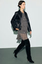 A person wearing a black leather jacket over a robe courte in mousseline fluide with a chic col chemise, paired with black knee-high boots and holding a black fringed bag, walks against a plain background.