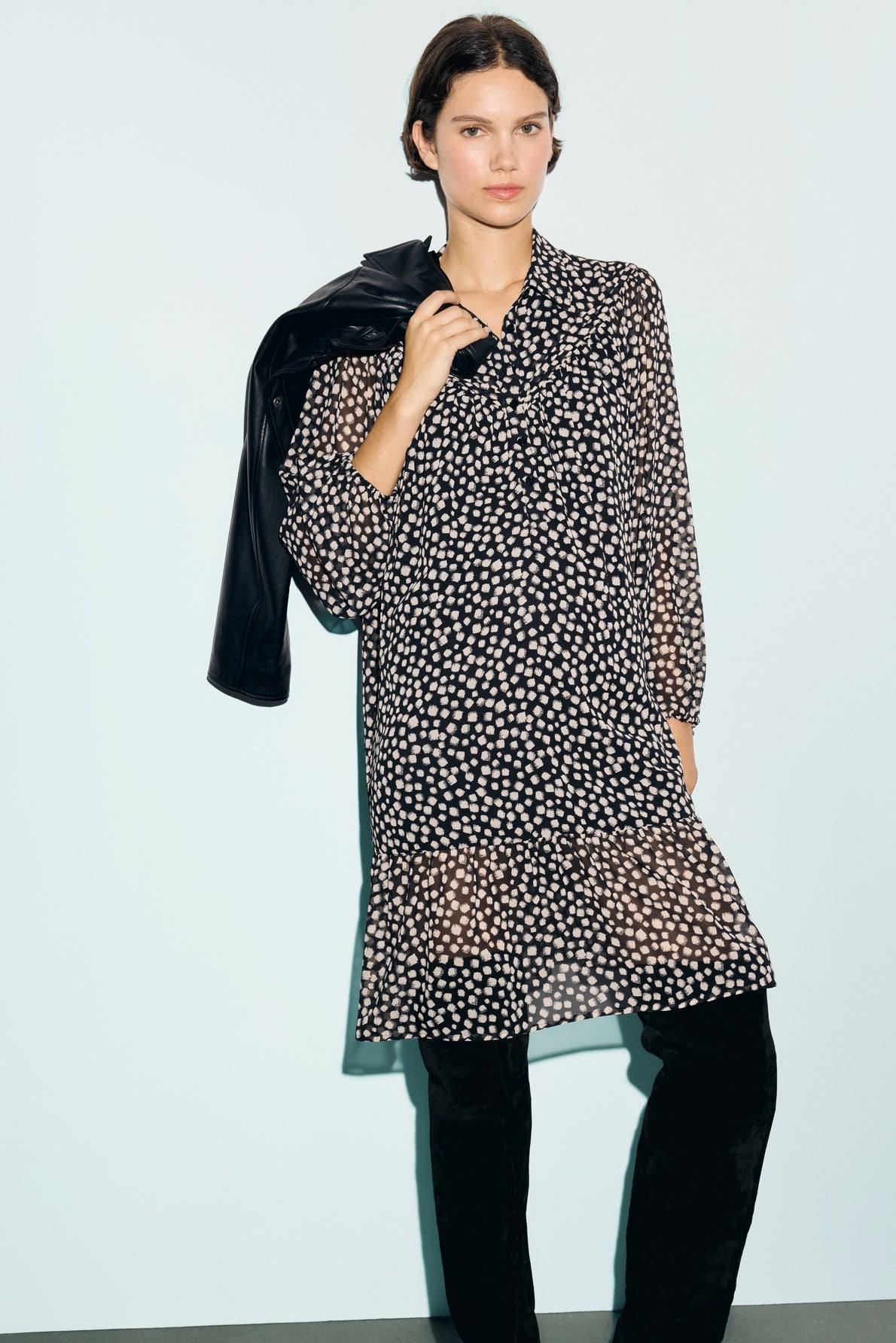 A woman wearing a black robe courte with white polka dots and black pants stands against a light background, the mousseline fluide fabric gently draping as she holds a black jacket over her shoulder. The chic col chemise detail adds an elegant touch to her look.