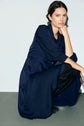 A woman in a bleu marine robe midi with rolled-up sleeves and a col chemise squats on the floor, resting her chin on her hand, looking at the camera against a plain background.