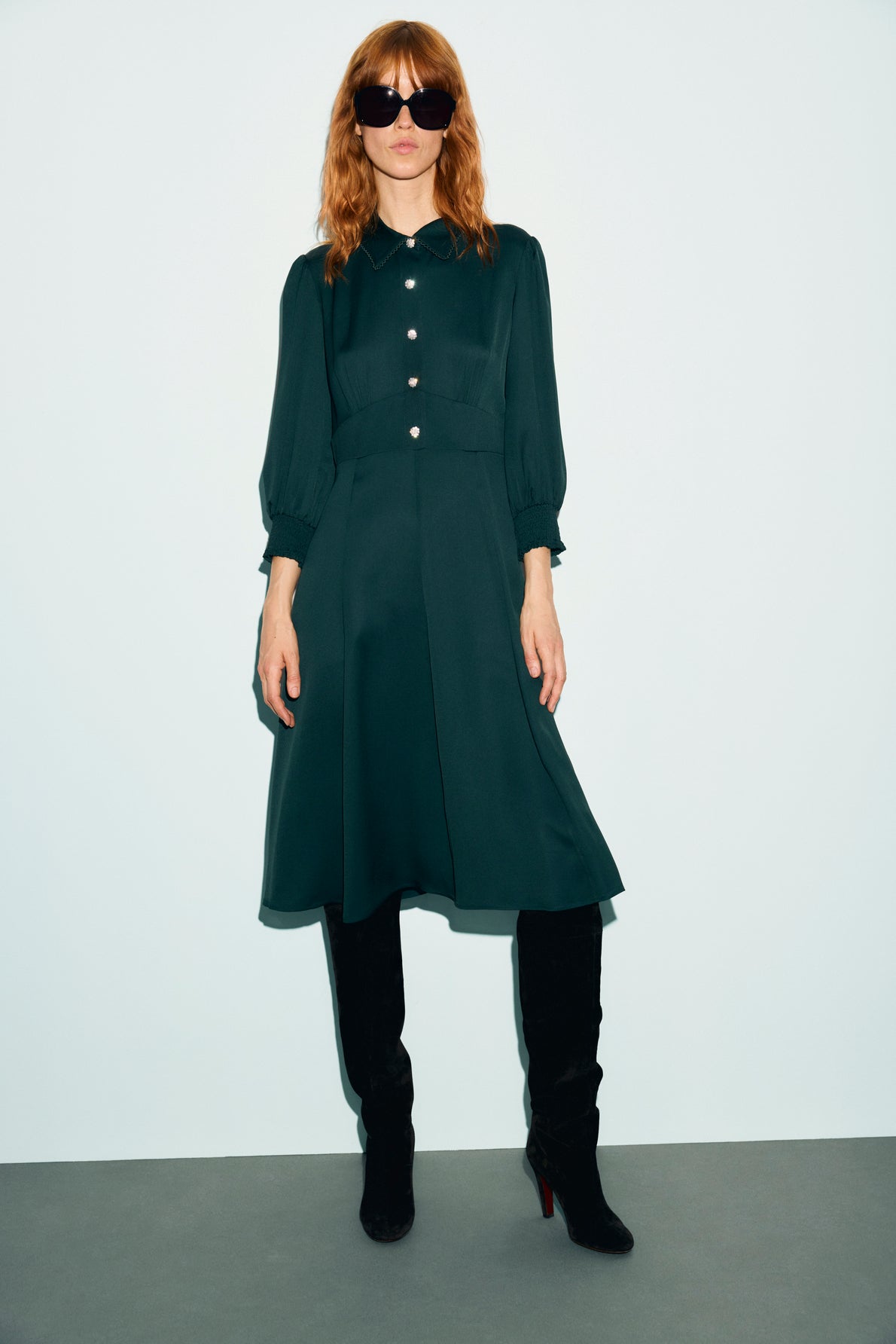 A woman with wavy red hair wears black sunglasses, a dark green robe midi à col chemise with buttons, and black knee-high boots, standing against a light background.