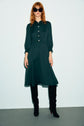 A woman with wavy red hair wears black sunglasses, a dark green robe midi à col chemise with buttons, and black knee-high boots, standing against a light background.