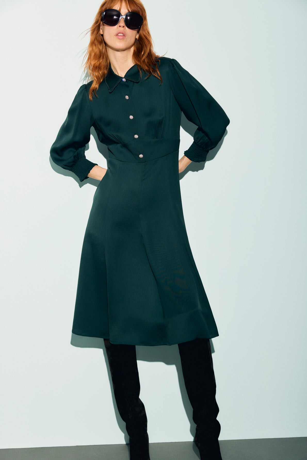A woman in sunglasses wears a dark green robe midi, long-sleeve, button-down with a col chemise and black knee-high boots, posing against a light background.