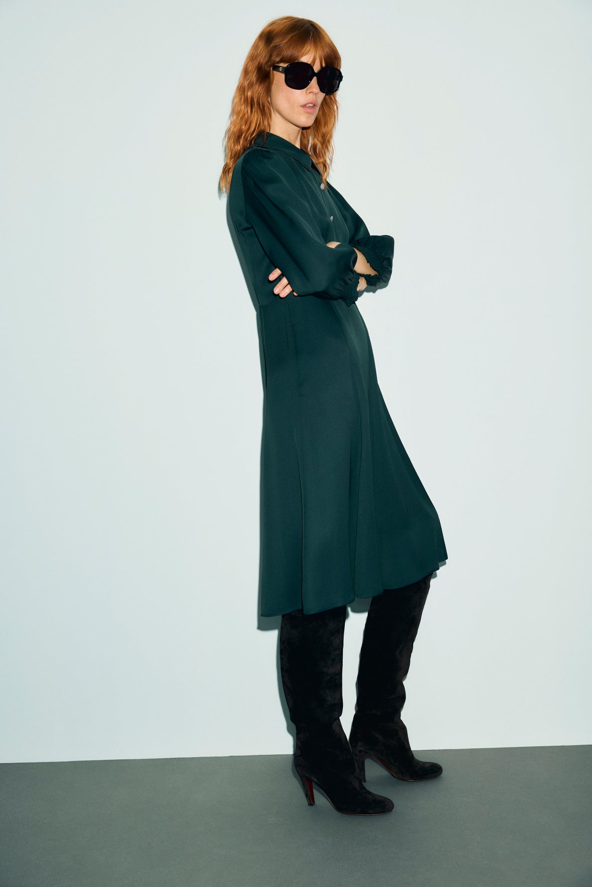 A woman with red hair wearing black sunglasses, a long dark green robe midi, and black knee-high boots stands with her arms crossed against a plain light-colored background.